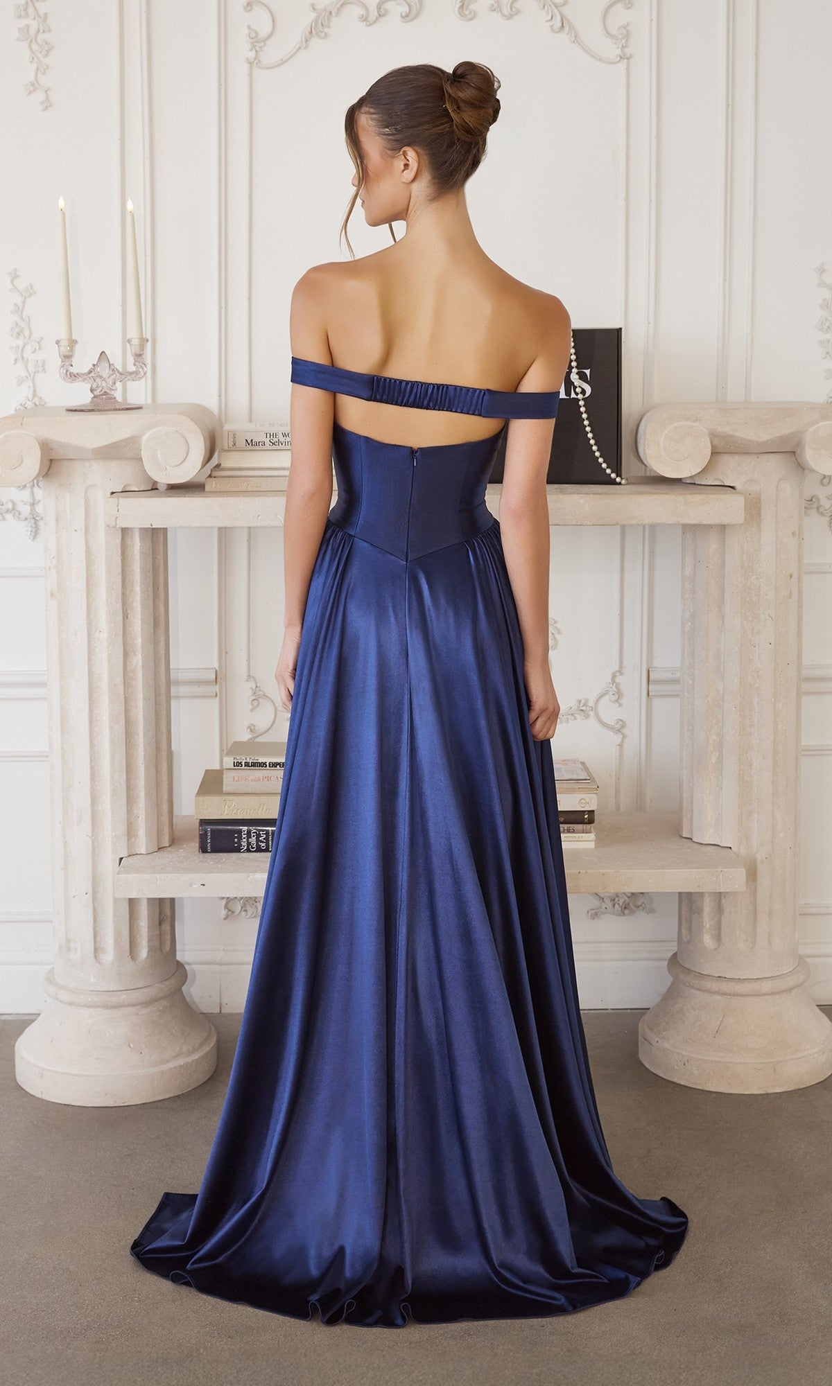A woman faces away in a floor-length, navy satin off-the-shoulder gown—Long Prom Dress: Amoris T821 by Cinderella Divine—standing before a white ornate mantel with books and candles, creating a timeless scene.